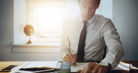 blog ERP Financial Management uomo in giacca e cravatta seduto alla scrivania e che scrive su un foglio