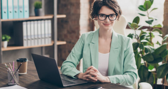 donna sorridente in giacca seduta alla scrivania di un ufficio con un portatile davanti dove è installato un ERP spedizioni